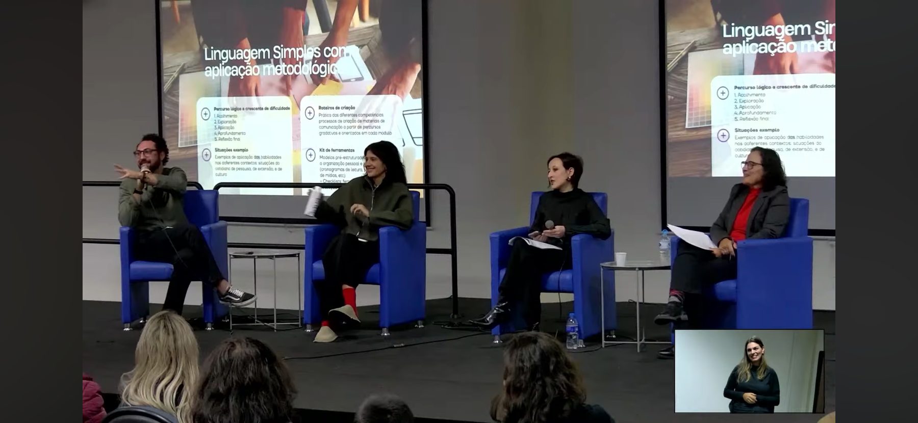 Mesa de debate com quatro pessoas em poltronas azuis diante de um telão sobre linguagem simples